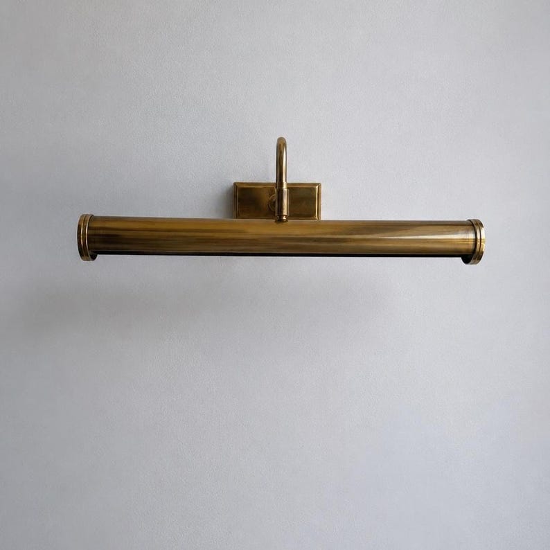 May include: A brass picture light fixture with a rectangular backplate and a curved arm. The light has a cylindrical shade and is designed to be mounted on a wall. The fixture is made of brass and has a warm, golden color.