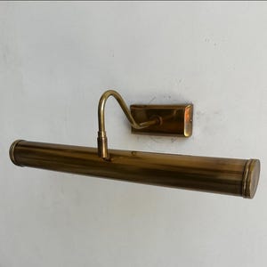 May include: A vintage brass picture light fixture. The fixture has a long, cylindrical light housing with rounded ends, a curved arm, and a rectangular wall mount. The brass finish gives it a classic, antique look.
