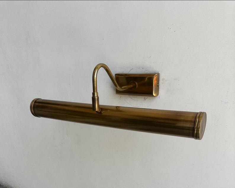 May include: A vintage brass picture light fixture. The fixture has a long, cylindrical light housing with rounded ends, a curved arm, and a rectangular wall mount. The brass finish gives it a classic, antique look.