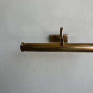 May include: A brass picture light fixture. The fixture has a long, cylindrical light housing with a rounded end. It is mounted on a rectangular backplate with a curved arm. The finish is a warm, golden color.