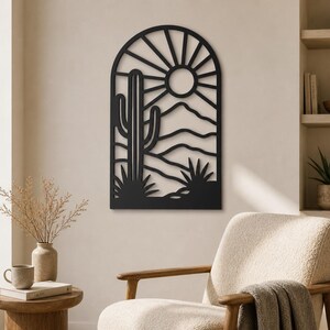 Op de afbeelding: Een zwart metalen wanddecoratie met een boogvormig frame en een woestijnlandschapontwerp. Het uitgesneden silhouet toont een hoge saguaro-cactus, glooiende heuvels en een zon met stralen die zich in de bovenste boog van het object bevinden.