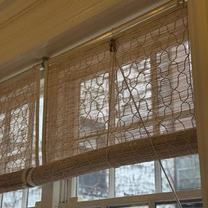May include: Beige bamboo window blinds, partially lowered, with a woven pattern. The blinds are mounted inside a white window frame, with a cord for adjusting the height. The window reveals a view of a building and trees.