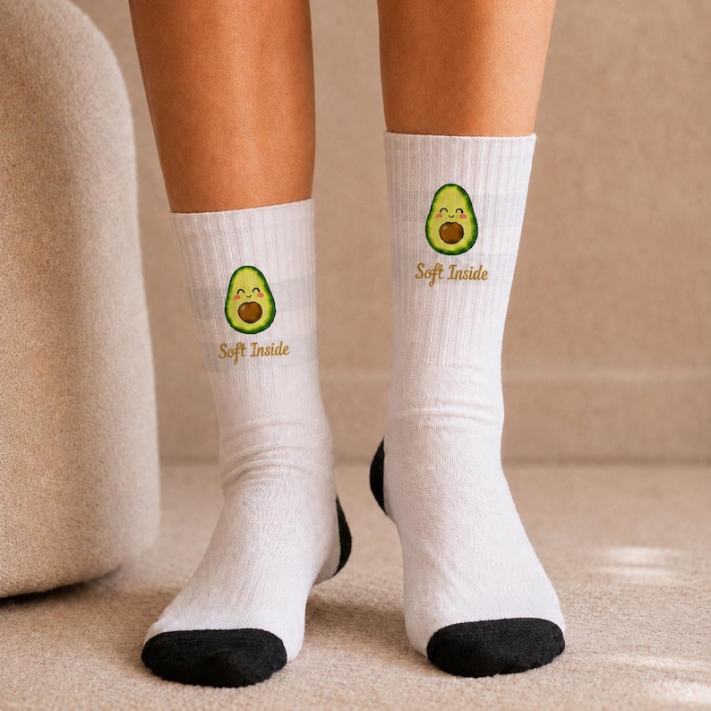 May include: White crew socks with a black toe and heel section. Each sock features a small, smiling avocado illustration centered on the shin area, accompanied by the text "Soft Inside" printed in a brown, cursive font below the avocado graphic.