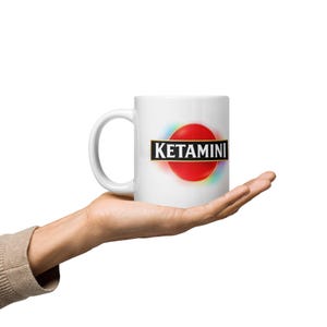 May include: White ceramic mug with the word "KETAMINI" in black capital letters on a black banner, set against a red circle with a rainbow gradient. The mug is held in a hand.