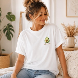 May include: A white short-sleeved t-shirt featuring a small graphic on the left chest of a smiling avocado with a heart-shaped pit. Below the avocado, the text soft inside is printed in a brown, cursive font. The shirt has a crew neckline.