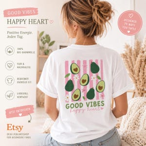 May include: A white t-shirt featuring a back graphic of pink vertical stripes, green avocados with heart-shaped pits, and small white flowers. The text GOOD VIBES and happy heart are printed in green and pink below the avocado illustration.