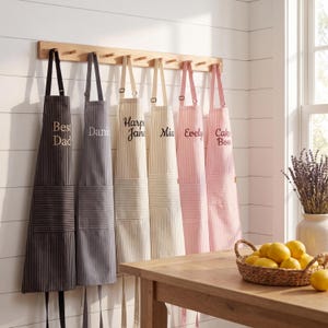 Op de afbeelding: Een verzameling gestreepte schorten die aan een houten rek hangen. De schorten zijn in verschillende kleuren, waaronder zwart, grijs, beige en roze, en voorzien van gepersonaliseerde tekst zoals "Best Dad" en namen. Een houten tafel met een mandje citroenen staat op de voorgrond.