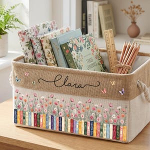 Puede incluir: Una cesta de almacenamiento rectangular de tela con una banda superior de arpillera color canela y una sección inferior crema. La cesta presenta un diseño impreso de flores silvestres coloridas y libros, con el nombre Clara en letra negra.