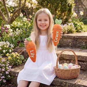 May include: A girl in a white dress holds two orange carrot-shaped plush toys, each with a name printed on them. A wicker basket filled with pastel-coloured Easter eggs sits nearby. The scene is set outdoors with a garden background.