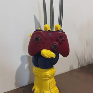 Peut inclure: Un support de manette imprimé en 3D en forme de main gantée jaune et bleue avec trois longues griffes argentées pointant vers le haut. La main tient une manette de jeu sans fil rouge foncé avec des joysticks et des boutons noirs, incluant un logo Xbox.