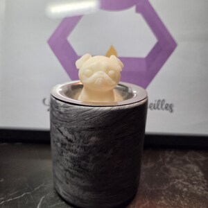 Puede incluir: Un pequeño derrite de cera con forma de perro pug de color crema se encuentra dentro de un calentador de cera cilíndrico, gris y negro veteado. El calentador tiene un borde plateado. Una imagen enmarcada con un gráfico de vela púrpura está en el fondo.