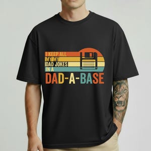 Op de afbeelding: Zwart t-shirt met een retro ontwerp met een diskette en de tekst "I KEEP ALL MY DAD JOKES IN A DAD-A-BASE" in oranje, geel en blauw. Het ontwerp is geplaatst tegen een achtergrond in zonsondergangstijl.