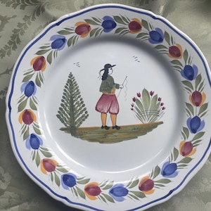 Op de afbeelding: Witte keramische plaat met een geschulpte rand, versierd met een handgeschilderde scène van een persoon die aan het vissen is. De rand heeft een blauw-wit ontwerp met kleurrijke bloemaccenten. De plaat heeft een diameter van ongeveer 25 cm.