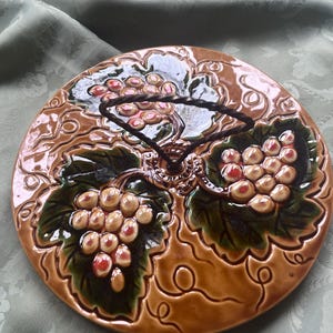 Op de afbeelding: Een ronde, bruine keramische plaat met een verhoogd druivenontwerp. De druiven zijn een mix van rode en crèmekleuren, met groene bladeren. De plaat heeft een decoratieve rand en een donkerbruin handvat.