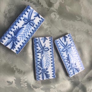 Può includere: Tre piastrelle rettangolari in ceramica con motivi floreali blu e bianchi. Ogni piastrella presenta un motivo floreale centrale con foglie circostanti e un bordo decorativo. Le piastrelle sono disposte su un tessuto verde chiaro.