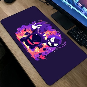 May include: A rectangular purple mousepad featuring a colorful graphic of cartoon ghost characters. The design includes purple, orange, and yellow elements. The mousepad is on a wooden desk, near a computer monitor and keyboard.