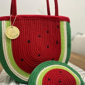 Puede incluir: Un bolso tote y un monedero a juego con temática de sandía. El bolso y el monedero están hechos de material tejido rojo, verde y blanco, con detalles de semillas negras. El bolso tote tiene asas y ribetes rojos.