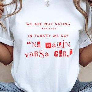 May include: A white short-sleeved t-shirt featuring red text that reads: WE ARE NOT SAYING "WHATEVER" IN TURKEY WE SAY "Ne HaLiN vaRsa GöR.". The text uses a mix of standard and ransom-note style lettering with some letters inside red boxes.