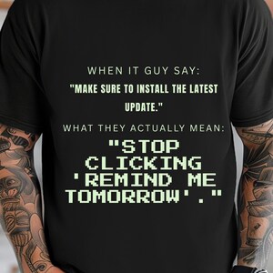 May include: A black short-sleeved t-shirt featuring white text that reads: WHEN IT GUY SAY: "MAKE SURE TO INSTALL THE LATEST UPDATE." WHAT THEY ACTUALLY MEAN: "STOP CLICKING 'REMIND ME TOMORROW'." The text is printed in a mix of standard and pixelated fonts.