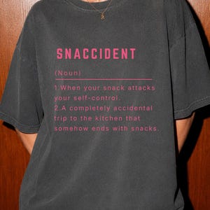 Op de afbeelding: Een antracietgrijs T-shirt met korte mouwen en roze tekst die luidt: SNACCIDENT (Noun) 1. When your snack attacks your self-control. 2. A completely accidental trip to the kitchen that somehow ends with snacks.