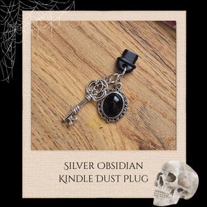 Könnte beinhalten: Ein silberner Kindle-Staubschutz mit einem Anhänger aus schwarzem Obsidianstein. Der Anhänger ist von einem verzierten, dekorativen Rahmen eingefasst. Der Stecker hat auch einen kleinen Zylinder-Anhänger und einen Schlüsselanhänger. Der Text "SILVER OBSIDIAN KINDLE DUST PLUG" steht darunter.