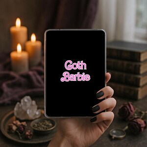 May include: A rectangular black tablet case featuring the text Goth Barbie in a stylized pink font with a white outline. The case has a clear border and is held in a hand against a dark, atmospheric background with candles and books.