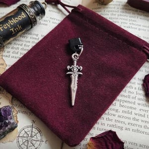 May include: A silver-toned metal dagger charm featuring a detailed hilt with curved crossguards and a pointed blade, attached to a small black plastic bead by a silver jump ring, resting on a deep burgundy velvet drawstring pouch.