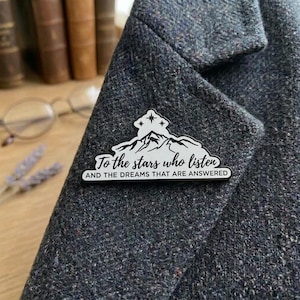 May include: A white enamel pin featuring a black mountain silhouette and three stars at the top. The text reads: To the stars who listen AND THE DREAMS THAT ARE ANSWERED. The pin is attached to a dark grey textured wool blazer fabric.