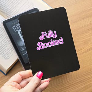 May include: A rectangular black card with rounded corners featuring the text Fully Booked in a stylized pink font with a white outline. The card is held in front of a digital e-reader device and an open book with visible printed text on the pages.