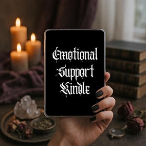 May include: A black e-reader case featuring the white text "Emotional Support Kindle" in a gothic-style font. The rectangular device has a clear protective shell and is held upright, displaying the bold lettering clearly against the dark background.