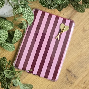 Puede incluir: Una funda de teléfono rectangular con rayas verticales en tonos alternos de burdeos oscuro y rosa claro. Un dije de metal dorado en forma de corazón con una letra A colgante está sujeto en la esquina superior derecha de la funda.