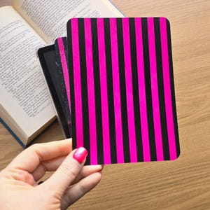 May include: A rectangular card featuring a bold pattern of vertical hot pink and black stripes. The card has rounded corners and is held in front of an open book with visible printed text on the pages, showcasing a high-contrast graphic design.