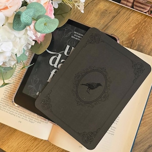 May include: A black e-reader case featuring an embossed oval frame with a raven illustration in the centre and decorative corner flourishes. The case rests on an open book with visible text, partially obscured by a cluster of white and pink flowers.