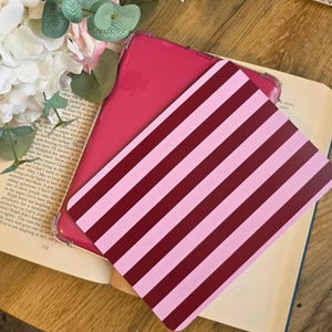 May include: A tablet case featuring a pattern of alternating dark red and light pink diagonal stripes. The case is positioned over a solid magenta tablet cover, which is resting on an open book with visible text and white floral accents.