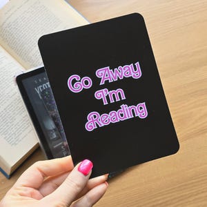 May include: A rectangular black sign with rounded corners featuring the text Go Away I'm Reading in a stylized pink font with a white outline. The lettering is centered on the surface, creating a bold contrast against the solid black background.