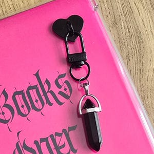 Peut inclure: Un pendentif en forme de cœur noir attaché à un fermoir mousqueton noir et une bélière en métal argenté retenant une pierre noire à facettes en forme de pointe. L'accessoire repose sur une surface rose vif avec des lettres gothiques noires.