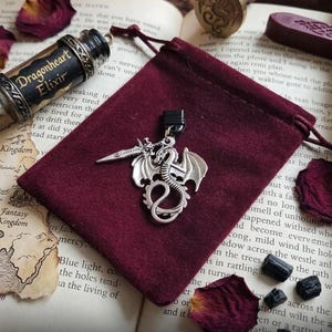 May include: A silver-toned metal pendant featuring a coiled dragon holding a sword, attached to a black cord bail. The pendant rests on a deep burgundy velvet drawstring pouch, accompanied by a glass vial labeled Dragonheart Elixir.