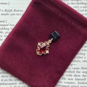 May include: A gold-toned pendant featuring a central pear-shaped red gemstone surrounded by intricate filigree metalwork and small clear stones. The pendant is attached to a small black rectangular display tab, showcasing a detailed ornate design.