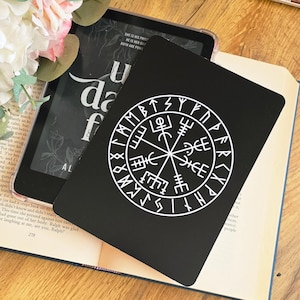 May include: A black rectangular e-reader cover featuring a white circular Vegvisir symbol surrounded by a ring of runic characters. The cover is positioned over an open book with visible text and a digital tablet displaying a book cover.