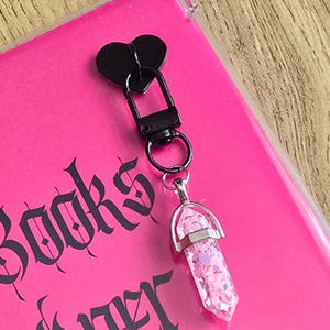 May include: A black heart-shaped clasp attached to a silver-toned metal bail holding a clear, hexagonal, crystal-like pendant filled with small pink and purple heart and star-shaped glitter pieces, resting on a bright pink surface with black text.
