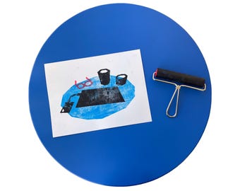 Puede incluir: Un papel blanco rectangular con una impresión en bloque de tinta azul de una escena de escritorio con gafas, dos tazas y un portátil, acompañado de un rodillo de goma negro con mango de metal plateado sobre una superficie azul circular.