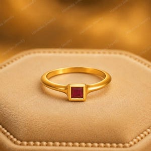 Op de afbeelding: Een gouden ring met een enkele vierkant geslepen dieprode edelsteen, gezet in een gladde, gepolijste gouden band. De ring heeft een eenvoudig en strak ontwerp waarbij de vierkante steen in een gladde zetting is geplaatst voor een contrast.