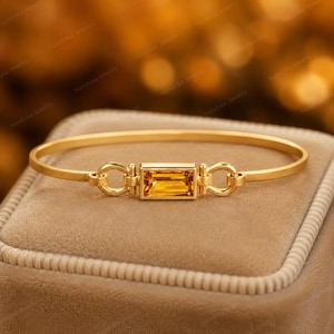 Può includere: Un bracciale rigido in oro con una pietra preziosa centrale di citrino giallo dal taglio baguette rettangolare. La pietra è incastonata in una montatura in oro collegata al cerchio da due piccoli anelli in oro su entrambi i lati.