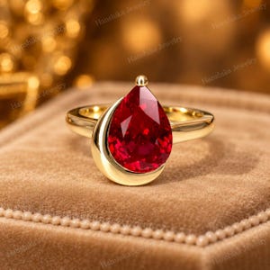 Puede incluir: Un anillo de oro con una gran piedra preciosa de color rojo intenso en forma de pera, montada en una banda de metal liso y pulido. El engaste rodea parcialmente la base de la piedra en forma de lágrima, creando un diseño elegante y moderno.