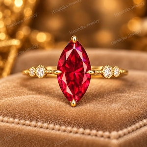 May include: A gold ring featuring a large marquise-cut red gemstone as the centre stone. The band is crafted from yellow gold and is accented on each side by two small, round, clear white stones set in a delicate, beaded metal design.