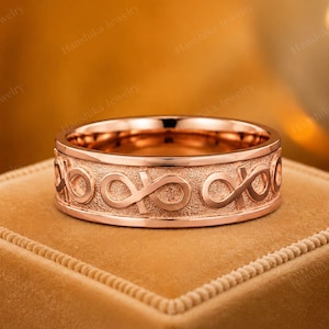 May include: A rose gold band ring featuring a repeating infinity symbol pattern embossed around the exterior. The centre band has a textured, matte finish, while the edges are polished and smooth, creating a contrast in the metal surface.