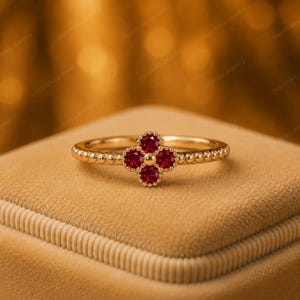 Könnte beinhalten: Ein Goldring mit einem vierblättrigen Blumenmotiv, das mit vier runden, tiefroten Rubinen besetzt ist. Die Ringschiene hat eine perlenartige Textur an den Außenkanten, und die Mitte der Blume ist mit einer kleinen, glatten Goldkugel akzentuiert.