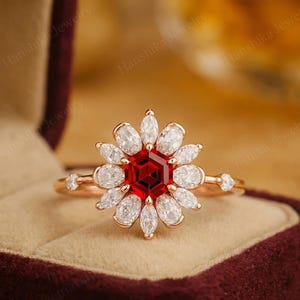 May include: A rose gold ring featuring a flower design. The center stone is a hexagonal, deep red gemstone, surrounded by white, petal-shaped stones. The band has small accent stones on each side.