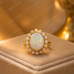 Könnte beinhalten: Ein Goldring mit einem großen, ovalen, schillernden Opal-Cabochon in der Mitte, umgeben von einem Kranz aus kleinen, runden weißen Perlen. Die Ringschiene besteht aus poliertem Gelbgold und bildet ein klassisches und elegantes Schmuckstück.