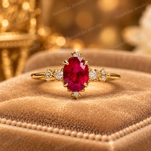 May include: A gold ring featuring a large, oval-cut, vibrant red gemstone at the centre. The setting includes smaller, round, clear accent stones arranged in clusters on either side of the central red stone, all mounted on a thin gold band.